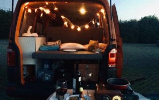 Interior de furgoneta camper con luces, transmite tranquilidad. En el plano delantero una mesa lista para cenar.