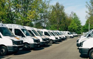 ¿Necesitas un seguro para tu flota de vehículos? Conjunto de furgonetas blancas de reparto aparcadas en batería en un entorno con árboles.